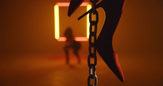 Woman wearing skin-tight clothes dancing in a dimly lit studio with a neon sign. Camera selective focus on the red high heels hanging from chains