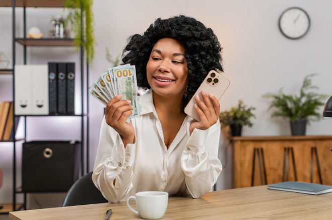 African American businesswoman surprised counts cash holding smartphone planning spending big profit