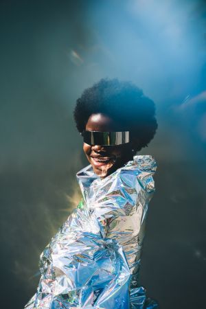 A futuristic portrait of a smiling African American woman wearing reflective visor sunglasses and wrapped in a metallic cloak.