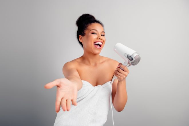 Smiling young woman in towel posing with hair dryer and extending hand on neutral background