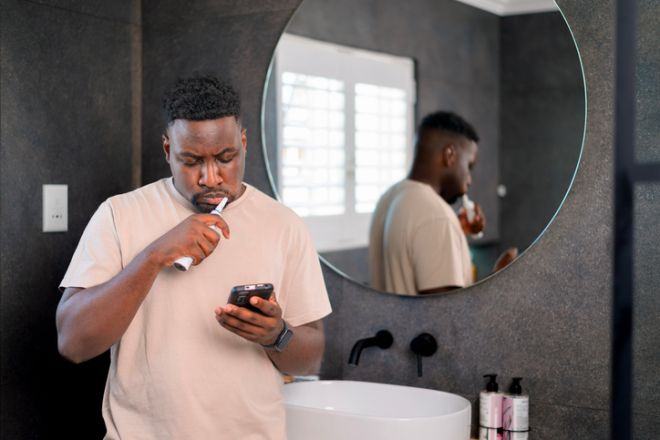 Man Using Smartphone While Brushing Teeth In Modern Bathroom Mirror Reflection