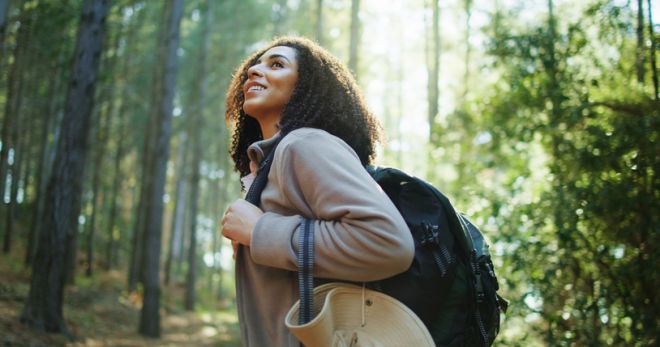 Thinking, hiking and happy woman in woods for adventure, eco tourism or holiday in nature. Vacation, backpacking and African person in forest for outdoor trip, travel and reflection for exploration