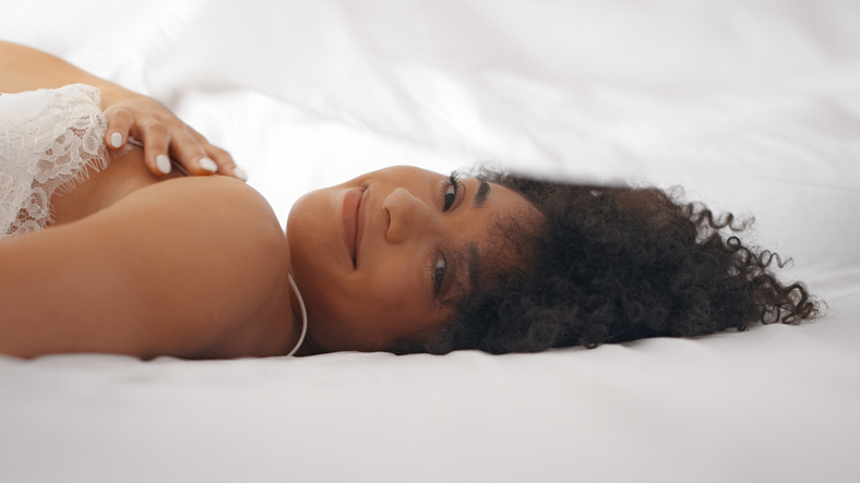 Beautiful young African ethnicity woman playfully rolling in the bed and hiding under white sheets. Looking at camera