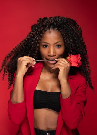 Sensual Hispanic woman with curly hair and rose in mouth posing in studio on red background