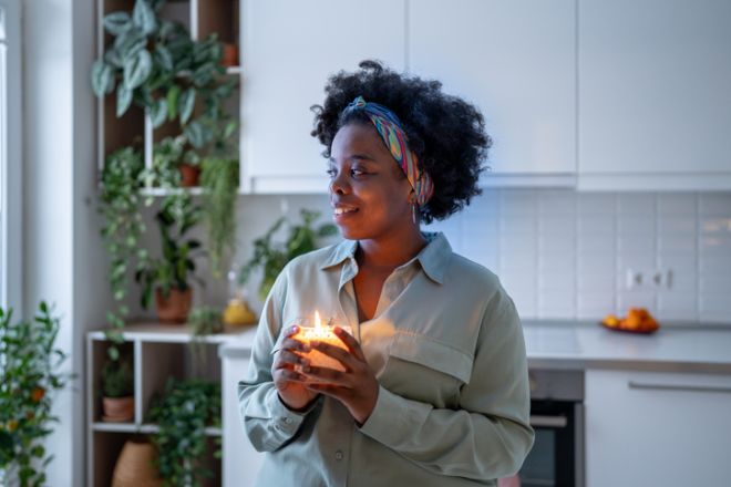 Serene pleased black woman lighting aroma candle feels gratitude enjoys cozy mood at home. Me time.