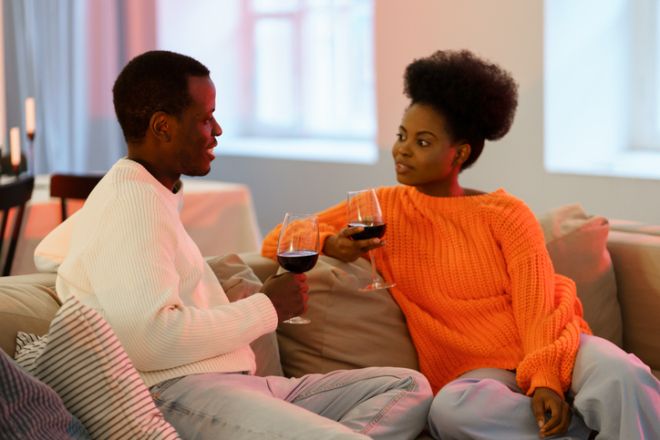 Happy african american couple man and woman drinking red wine at home, relaxing on sofa and talking