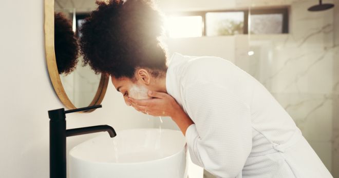 Woman, water and wash face for skincare in bathroom, acne control and cleansing routine at mirror. African person, dermatology and soap in home for facial treatment, skin hydration and exfoliation