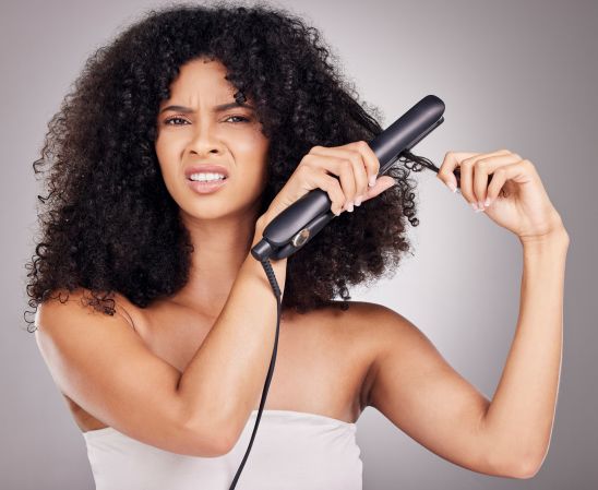 Sad, face portrait and woman with hair straightener in studio isolated on gray background. Haircare damage, hairloss and black female model angry with flat iron for hairstyle after salon treatment.
