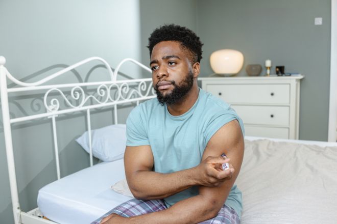 Man sitting on bed in bedroom injecting medication into his upper arm