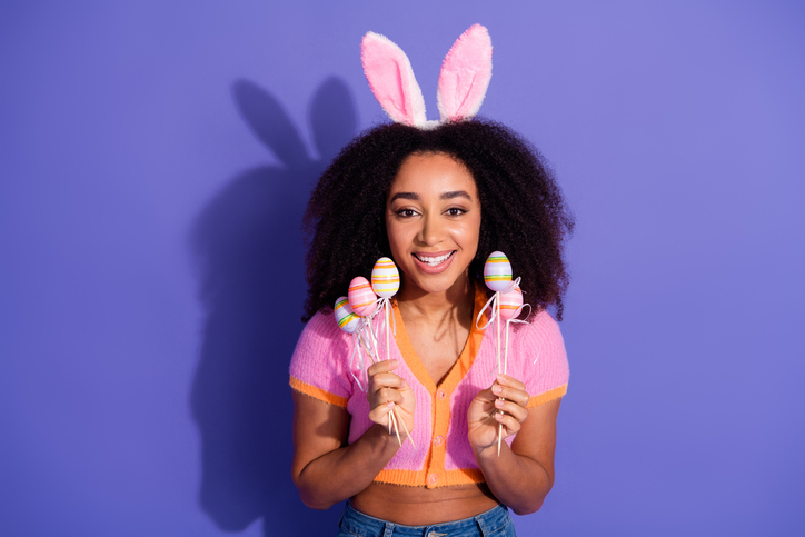 Smiling young woman with Easter bunny ears holding colorful egg decorations against a purple background celebrating spring and joy