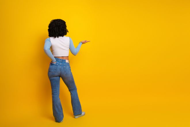 Back view of a stylish young woman pointing upwards against yellow background in casual wear for advertisement or presentation.