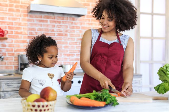 Mother slicing carrot while her daughter helping and playing beside her preparing fresh salad representing teamwork bonding healthy lifestyle family happiness at home