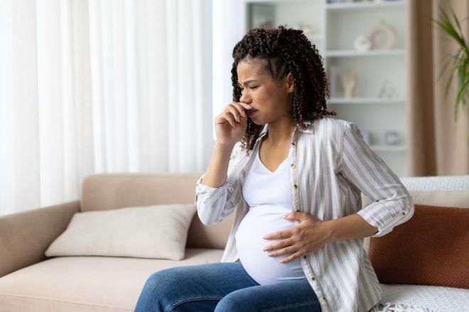 Pregnant African American woman sitting on sofa, covering mouth and looking unwell