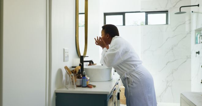 Black woman, makeup and morning by bathroom mirror, home or product for beauty with dermatology. African person, skincare and cosmetics in profile with touch for results, routine or wellness in house