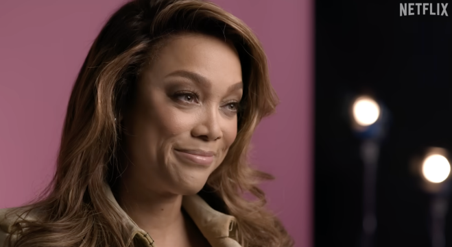 A woman with long, wavy brown hair smiling at the camera against a pink background, with the Netflix logo visible.