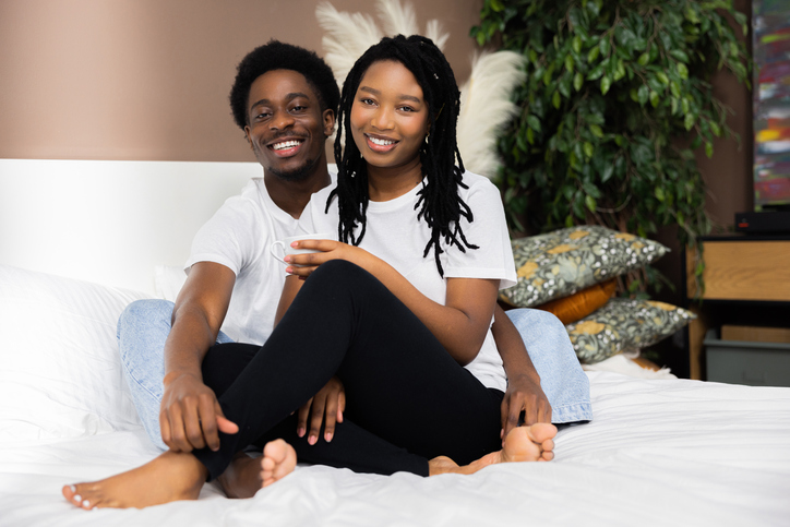 Picture of delighted smiley woman and man sitting on bed talking while drinking coffee in the morning smiling. Happy family relationship concept
