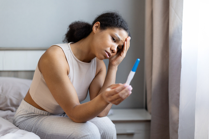 Young black woman sitting on bed holding pregnancy test with worried expression