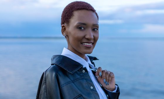 Young black woman with short dark hair in black leather jacket looking at camera with calm water and sky in background