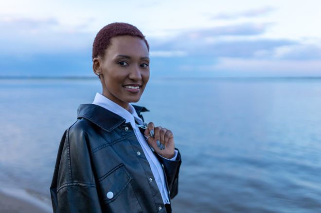 Young black woman with short dark hair in black leather jacket looking at camera with calm water and sky in background