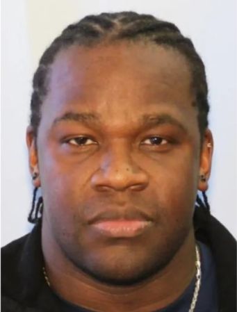 A close-up portrait of a young Black man with braided hair, wearing a black shirt and looking directly at the camera with a serious expression.