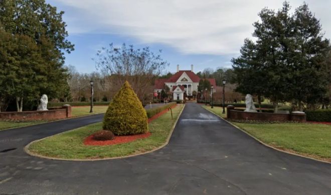 A long driveway leading to a large, red-roofed house surrounded by trees and landscaping in a park-like setting.