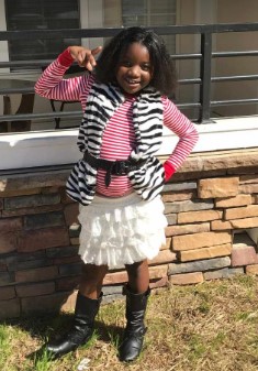 A smiling young girl wearing a striped shirt, white skirt, and black boots standing in front of a brick wall.