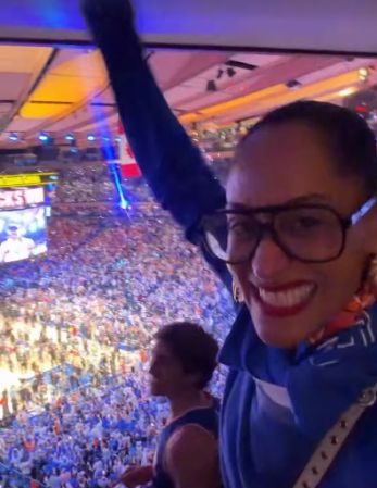 A smiling person in glasses looking out over a large, crowded arena or stadium filled with people.