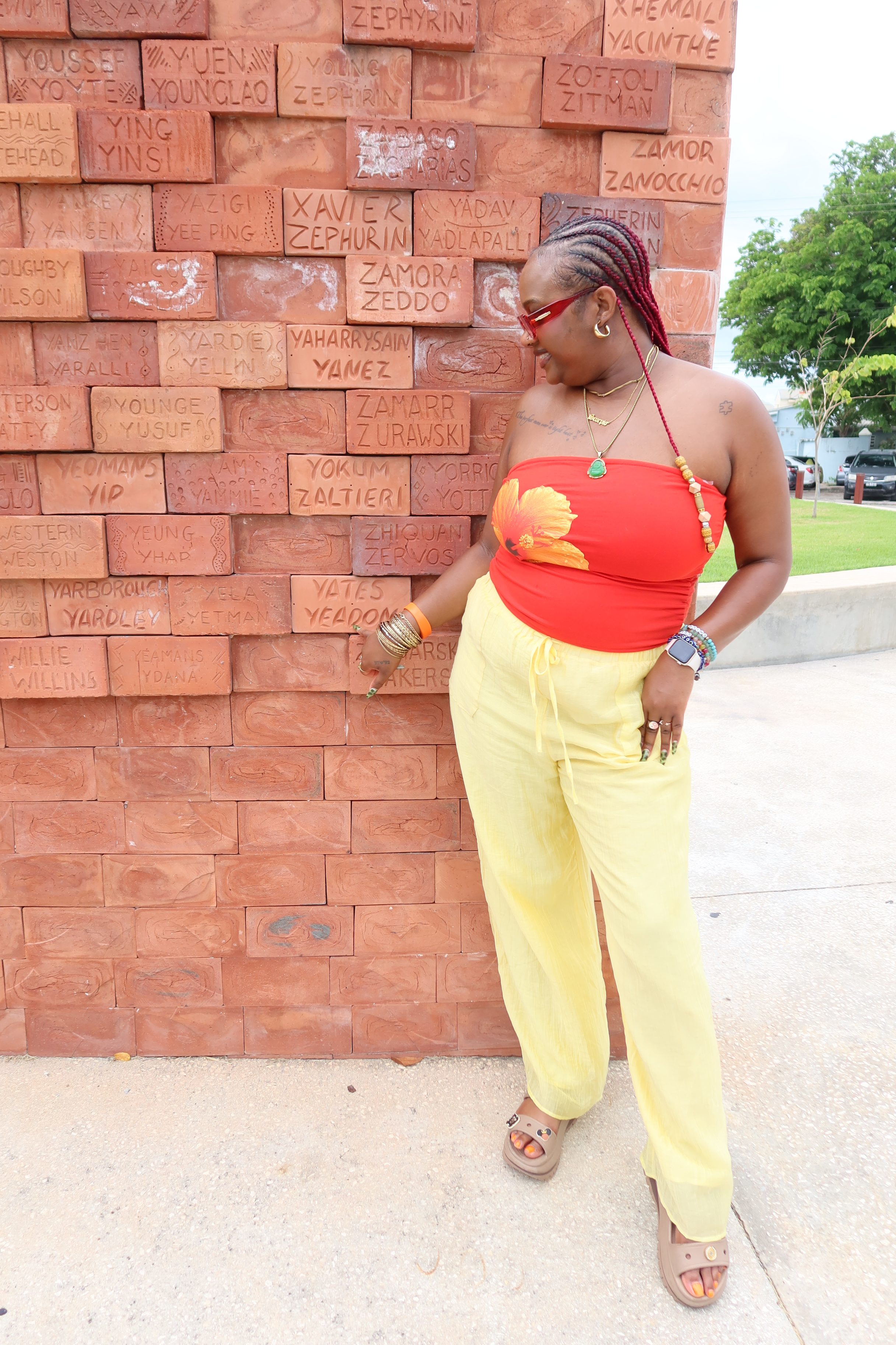 A woman wearing a red and yellow outfit stands in front of a brick wall with various names and words written on it.