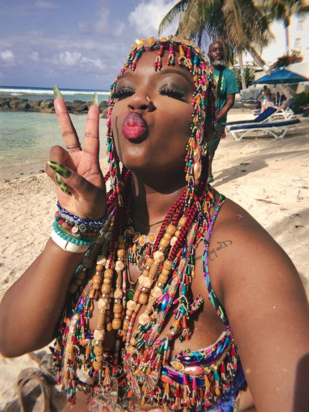 A person with colorful beaded hair and jewelry making a peace sign gesture on a beach with palm trees in the background.