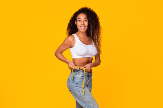 Black lady measuring her waistline with tape, smiling in studio