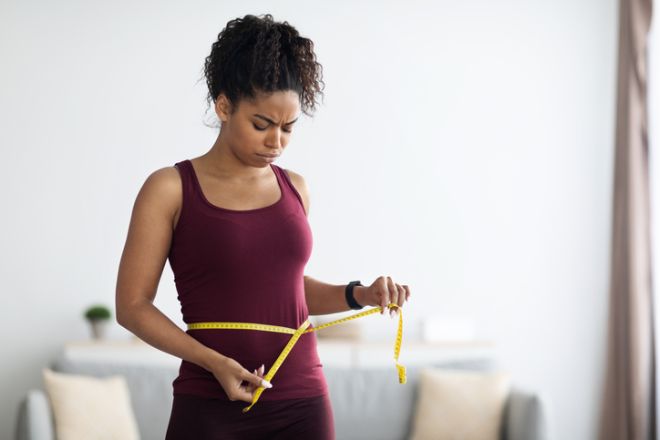 Unhappy african american woman in sportswear measuring waist