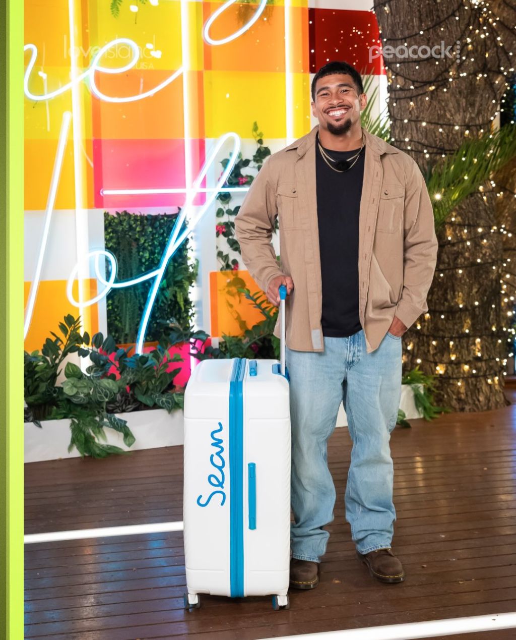 A smiling man standing in front of a colorful, neon-lit backdrop holding a Sloan-branded suitcase.