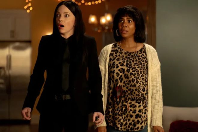 Two women, one wearing a black jacket and the other a leopard print top, standing in a dimly lit room with string lights.