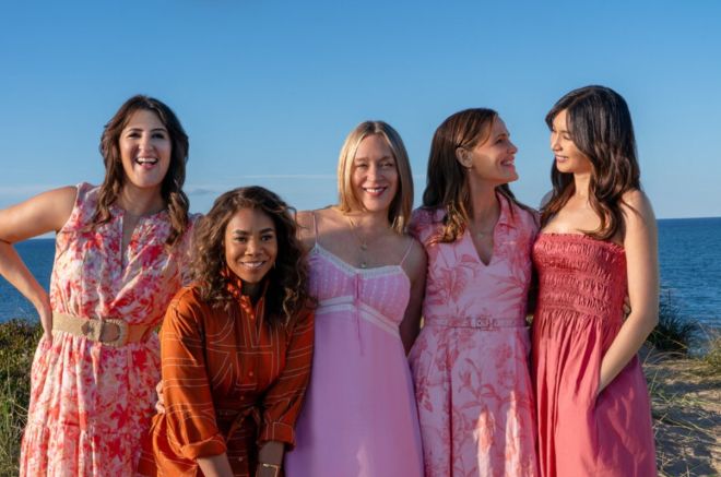 A group of five young women in colorful floral dresses standing together on a beach with the ocean in the background.