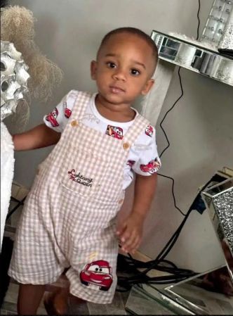 A young child wearing a white and red checkered outfit with cartoon car designs, standing in a room with various objects in the background.
