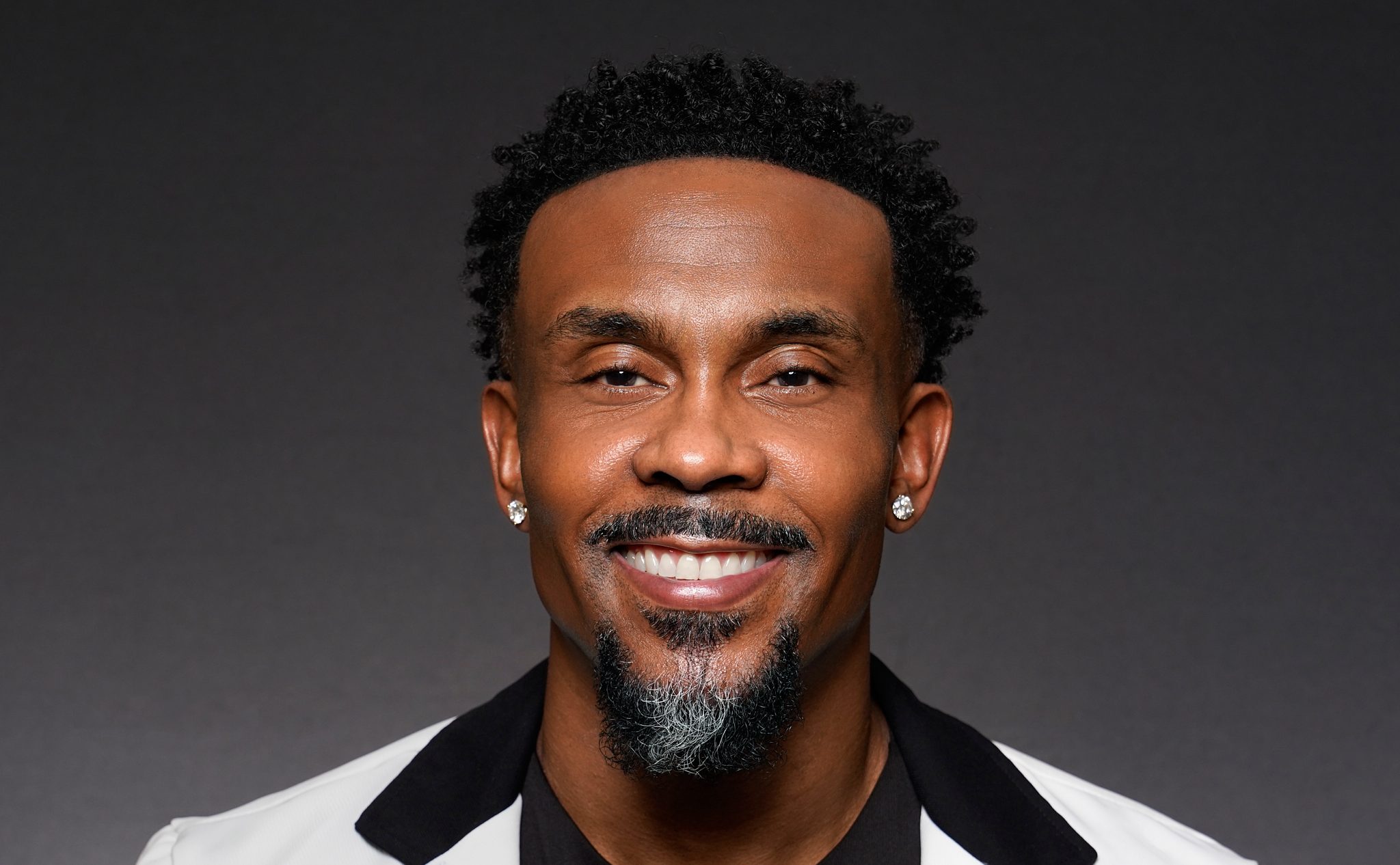 A smiling Black man with a beard wearing a white suit jacket and black shirt.