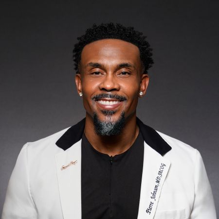 A smiling Black man with a beard wearing a white suit jacket and black shirt.