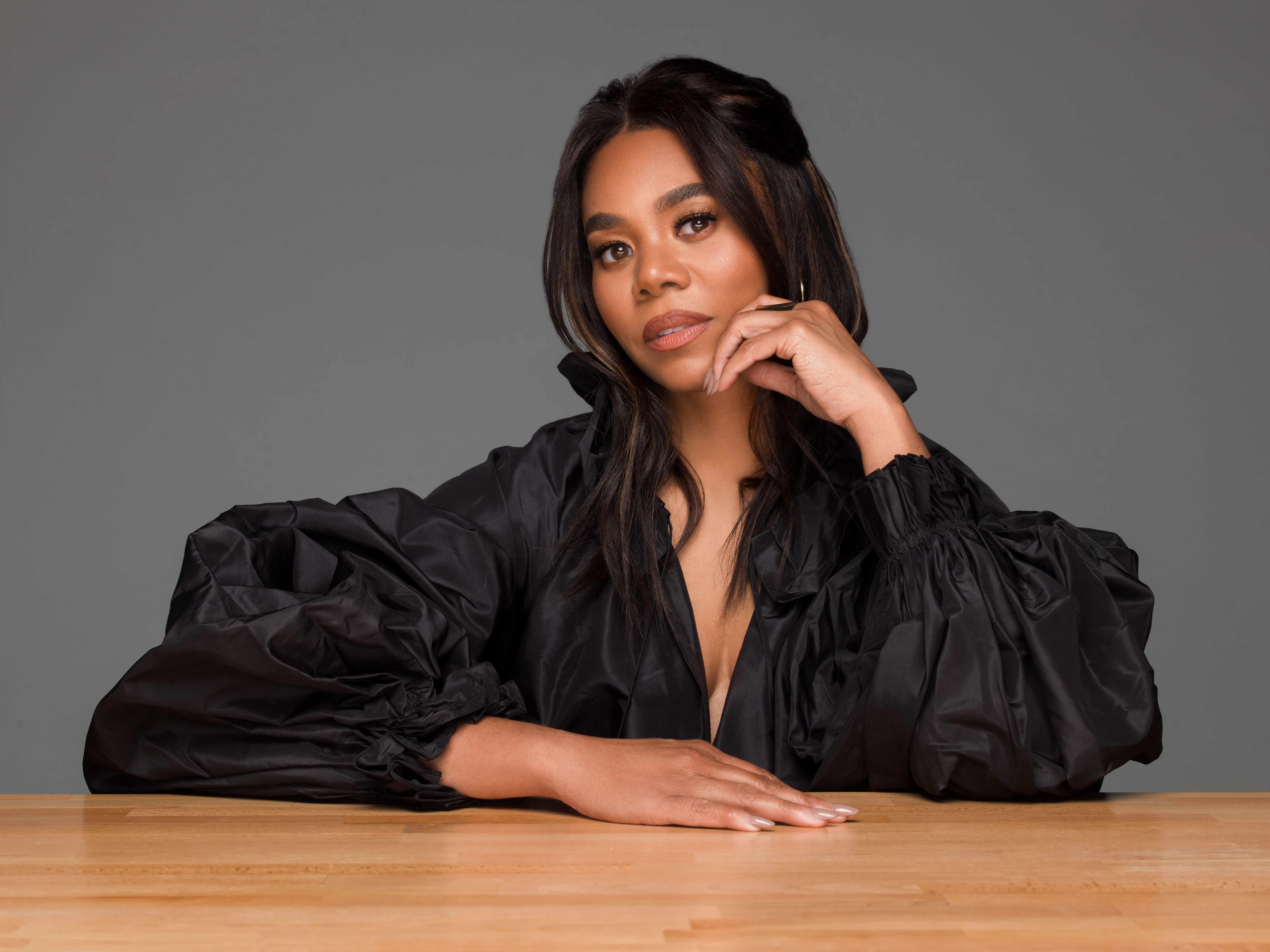 A woman with long dark hair wearing a black puff-sleeved dress sits at a wooden table, resting her chin on her hand with a thoughtful expression.