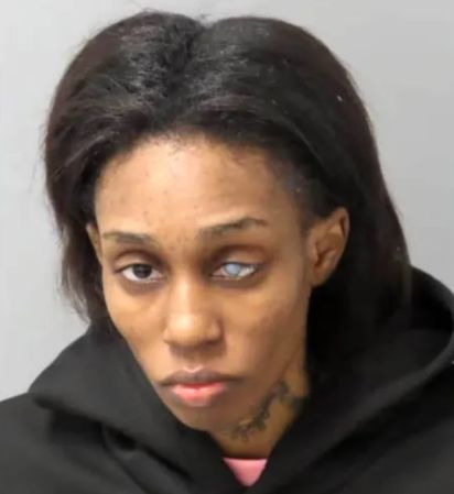 A close-up portrait of a serious-looking woman with dark hair and a tattoo on her neck, wearing a black hooded jacket.