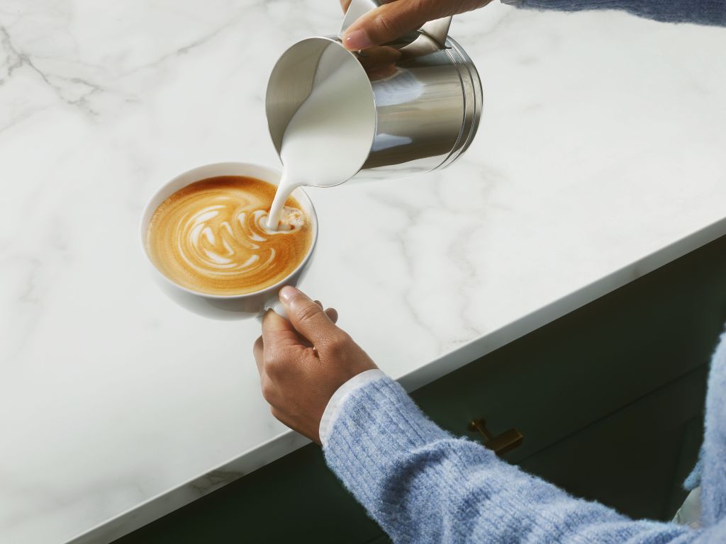 A person's hand pouring steamed milk into a cup of coffee, creating a latte art design on the surface.
