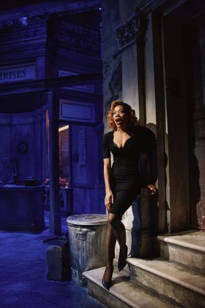 A woman in a black dress standing on steps in a dimly lit urban setting, with a trash can and signage visible in the background.