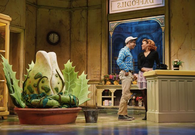 Two people, a man and a woman, standing and talking in an indoor setting with a large plant sculpture and a clock on the wall.