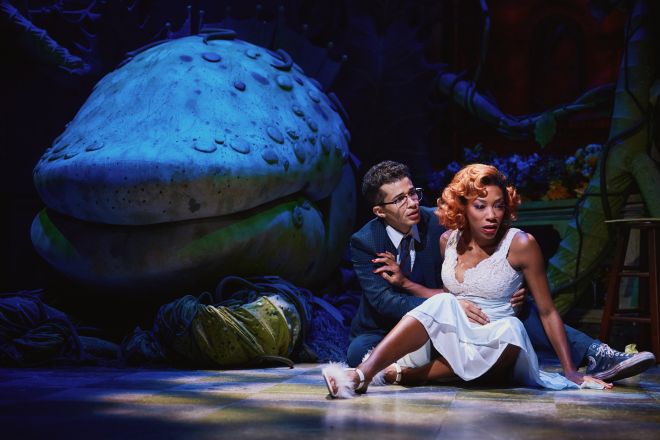 Two people, a man and a woman, sitting on the ground in a fantastical, blue-lit setting with a large, mushroom-like structure in the background.