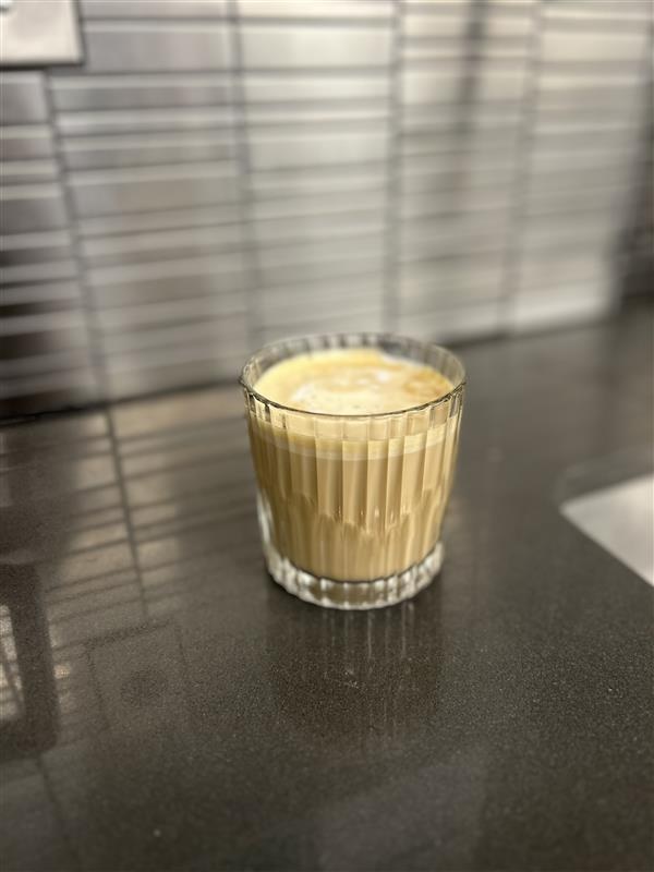 A glass filled with a creamy, frothy beverage on a dark surface, with a blurred background of shelves or racks.