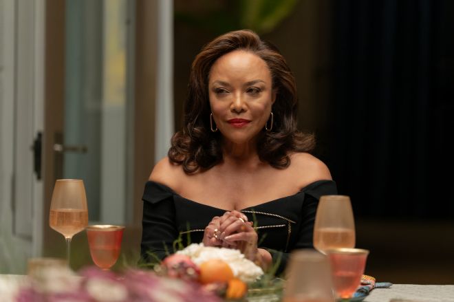 A woman with dark curly hair wearing a black off-the-shoulder dress sits at a table with wine glasses and a floral arrangement.