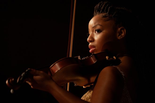 A young woman with braided hair holds a violin and gazes intensely, illuminated by a warm spotlight against a dark background.