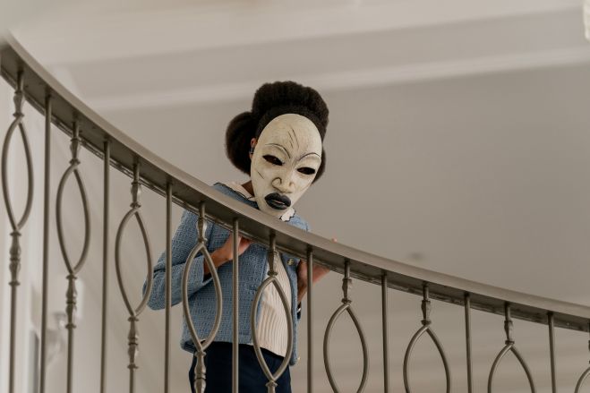A masked figure in a blue outfit peers through a metal railing.