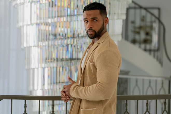 A man in a beige suit standing in a room with bookshelves in the background.