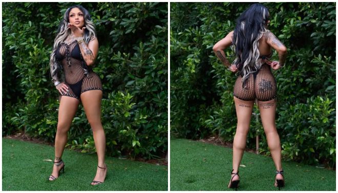 A woman wearing a black fishnet bodysuit and high heels stands in a lush, green outdoor setting.