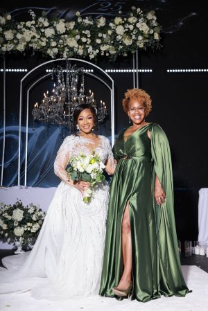 Two women in formal attire posing in front of a floral arch and chandelier at a wedding or formal event.